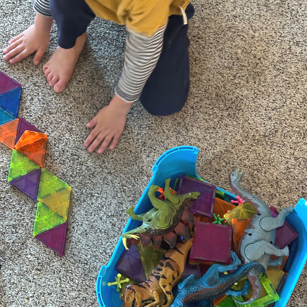 boy playing with magnet tiles and dinosaurs on carpet, distracting toddlers during homeschool lessons for 6 kids. 